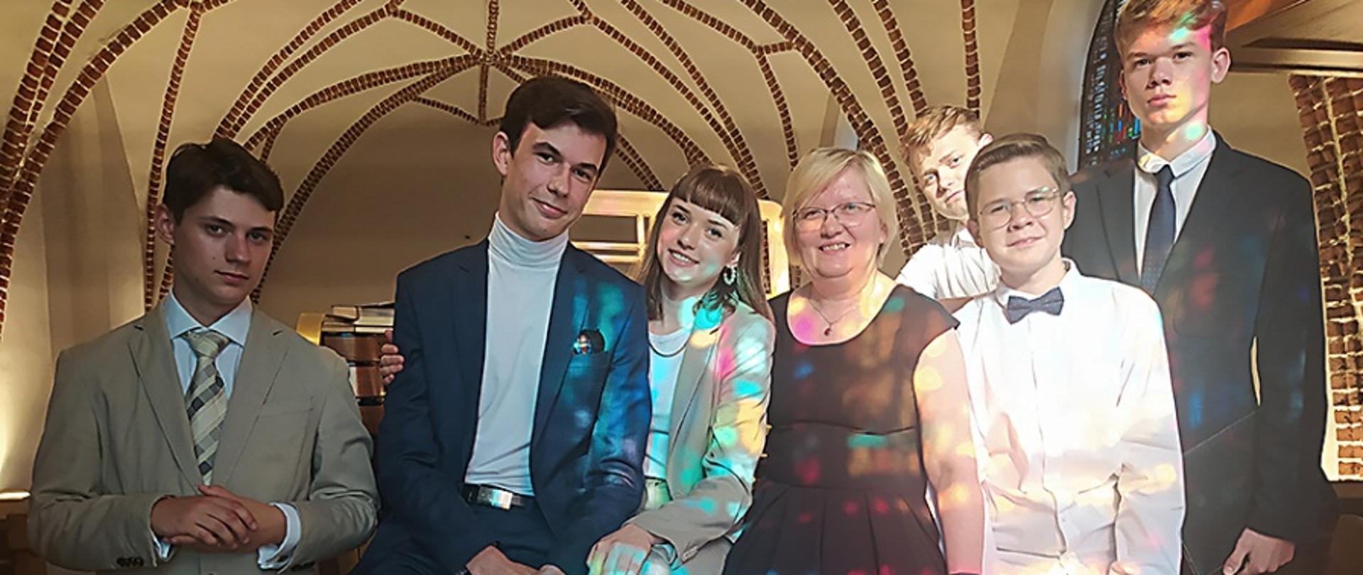 Muzycy w strojach galowych na chórze klasztoru, w tle sklepienie kościoła. Oświetleni kolorowym światłem witraża.