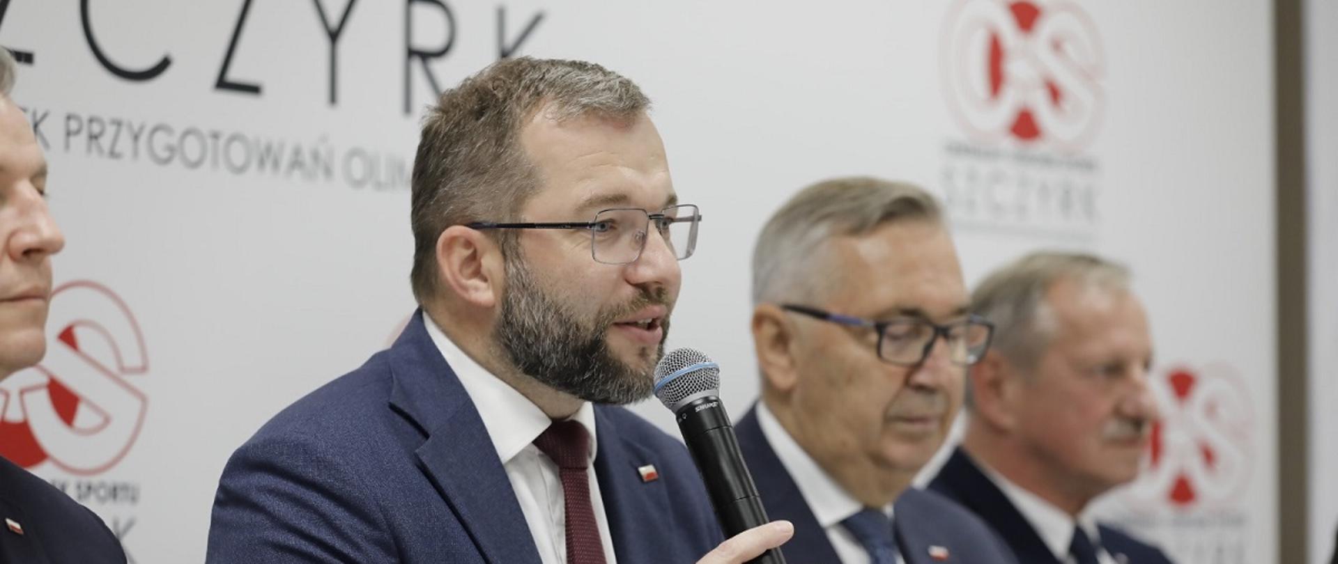 Minister funduszy i polityki regionalnej Grzegorz Puda siedzi przy stole i przemawia do mikrofonu, po jego prawej stronie wiceminister pracy i polityki społecznej Stanisław Szwed