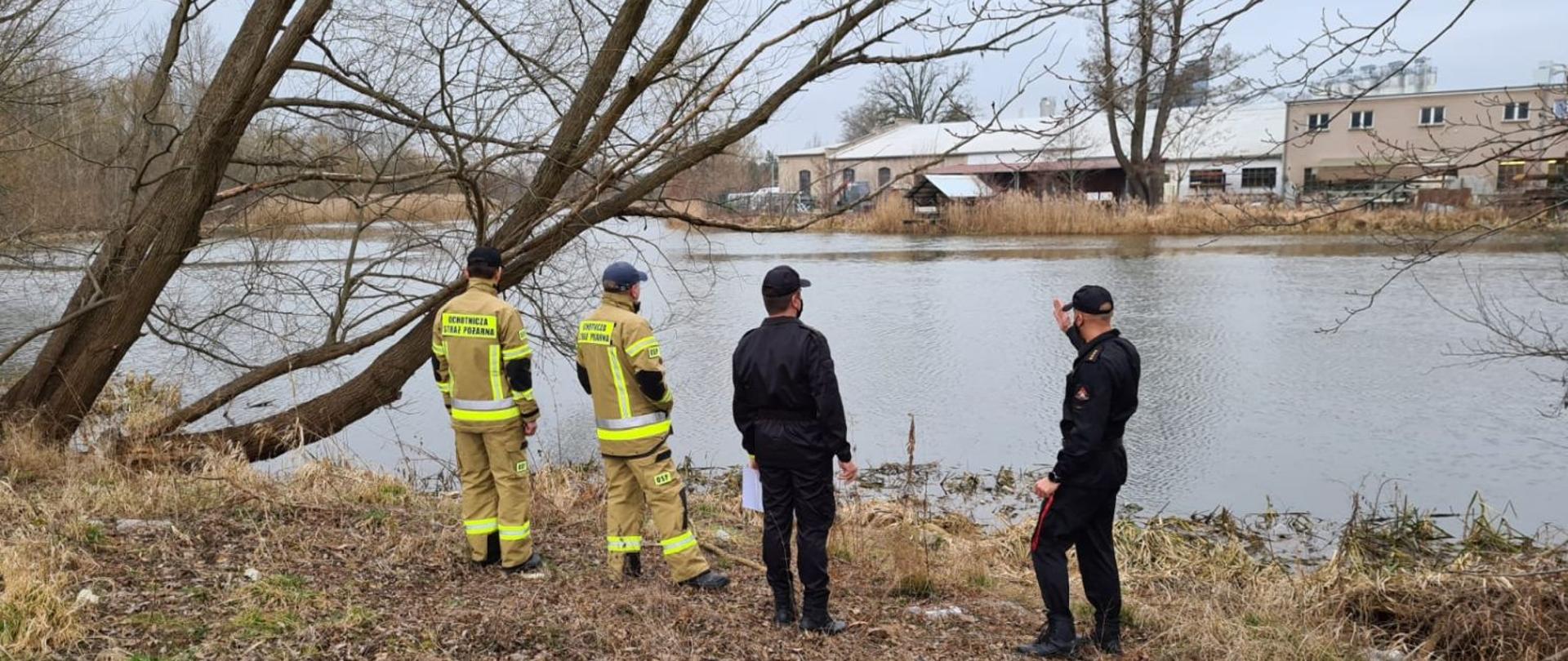 Czterech strażaków stojących na brzegu rzeki. Wszyscy obserwują stan wody i wałów przeciwpowodziowych. W tle widać wodę i budynki.