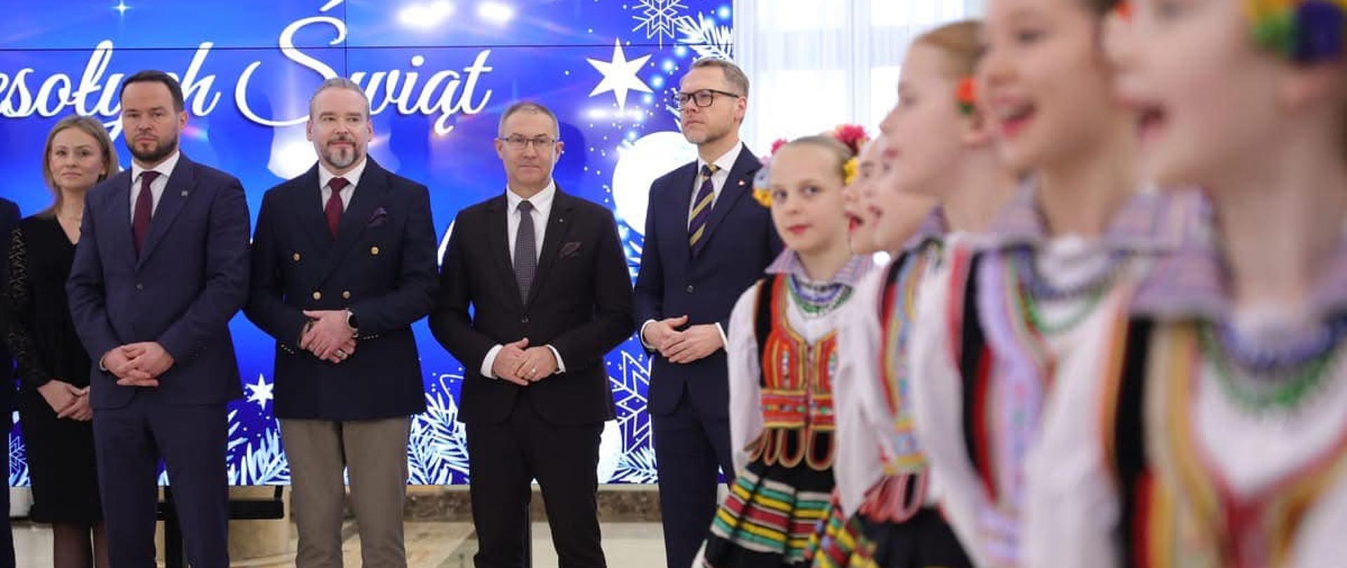 Czterech mężczyzn stojących na tle życzeń świątecznych, z prawej dziewczynki ubrane w stroje ludowe