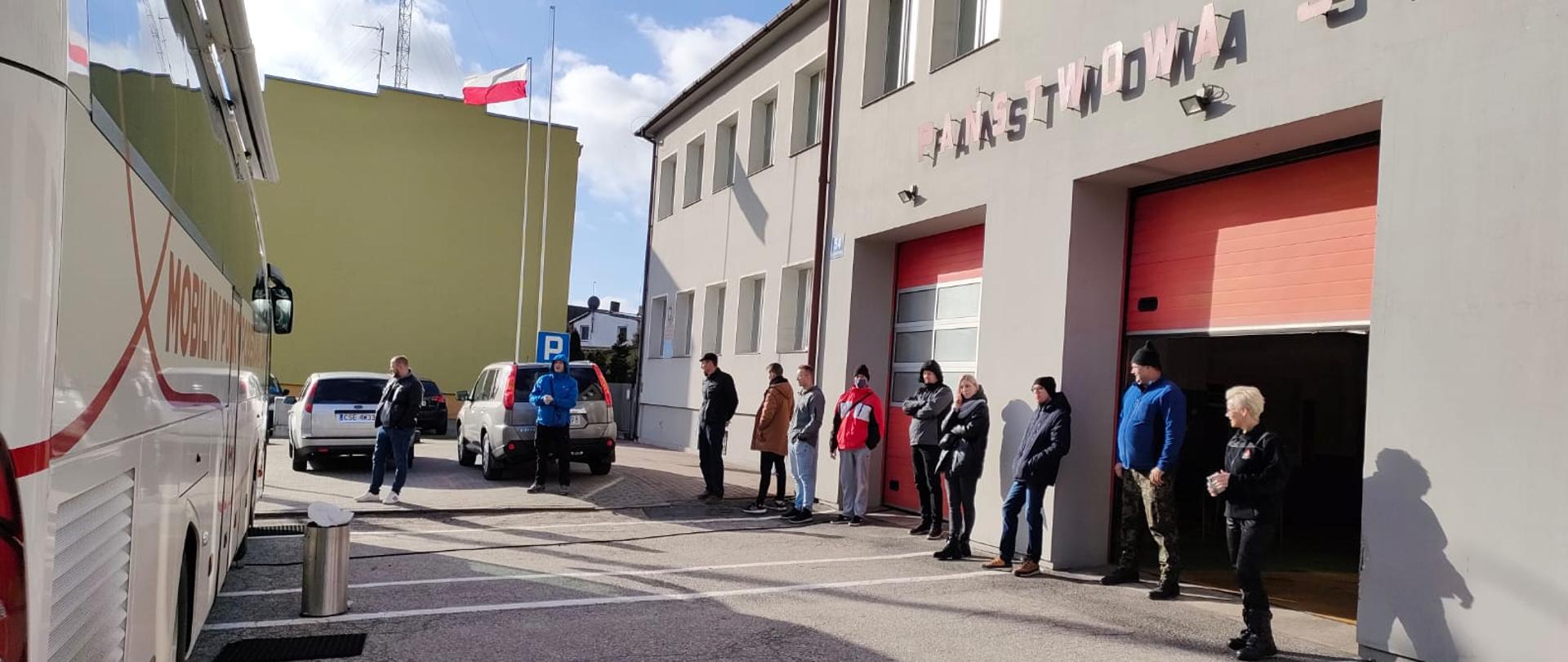 Zdjęcie wykonano w ciągu dnia przedstawia grupkę ludzi czekającą w kolejce do wejścia do Krwiobusa ustawionego przed Komendą Powiatową Państwowej Straży Pożarnej w Sępólnie Krajeńskim. Po lewej stronie Krwiobus, po prawej budynek Komendy.