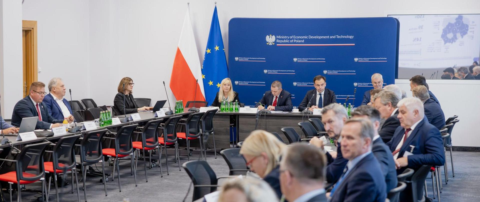 Widok na salę konferencyjną. Stoły ustawione w podkowę, po zewnętrznych stronach siedzą uczestniczy spotkania. U szczytu stołu delegacja MRiT z min. Domańskim. W tle flagi PL iUE, żsianka z logotypami MRIT, nad nią godło Polski.