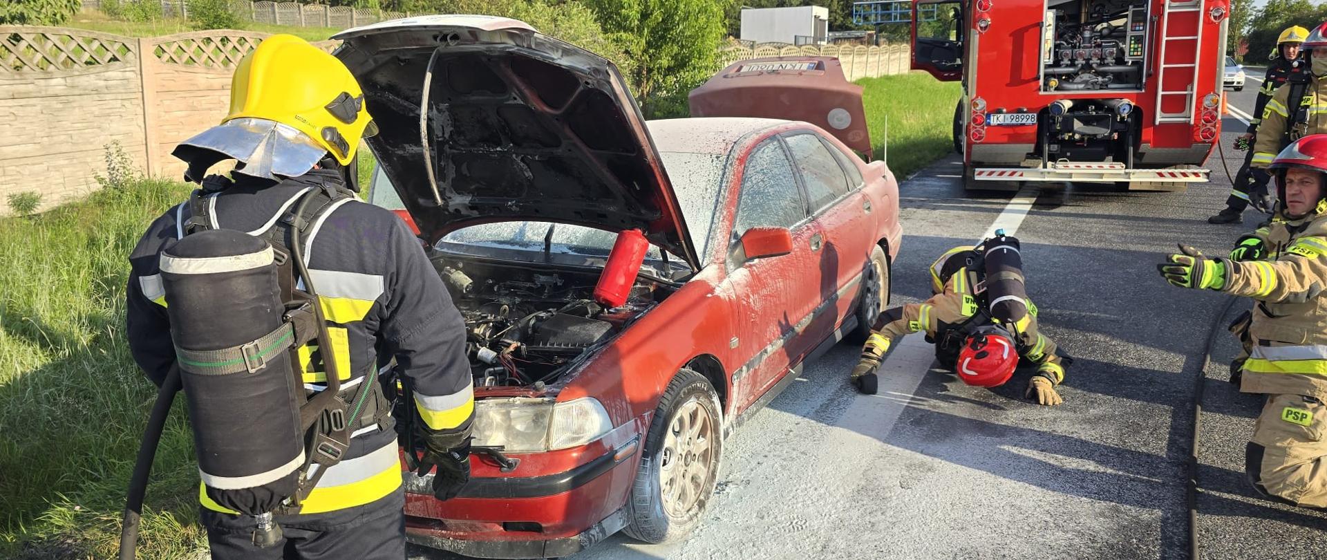 Zdjęcie przedstawia ratowników podczas akcji. Jeden z nich stoi przed otwartą maską spalonego pojazdu, drugi zagląda pod samochód potwierdzając brak zagrożenia, Auto i część jezdni zasypana jest proszkiem gaśniczym. W głębi stoi samochód pożarniczy.