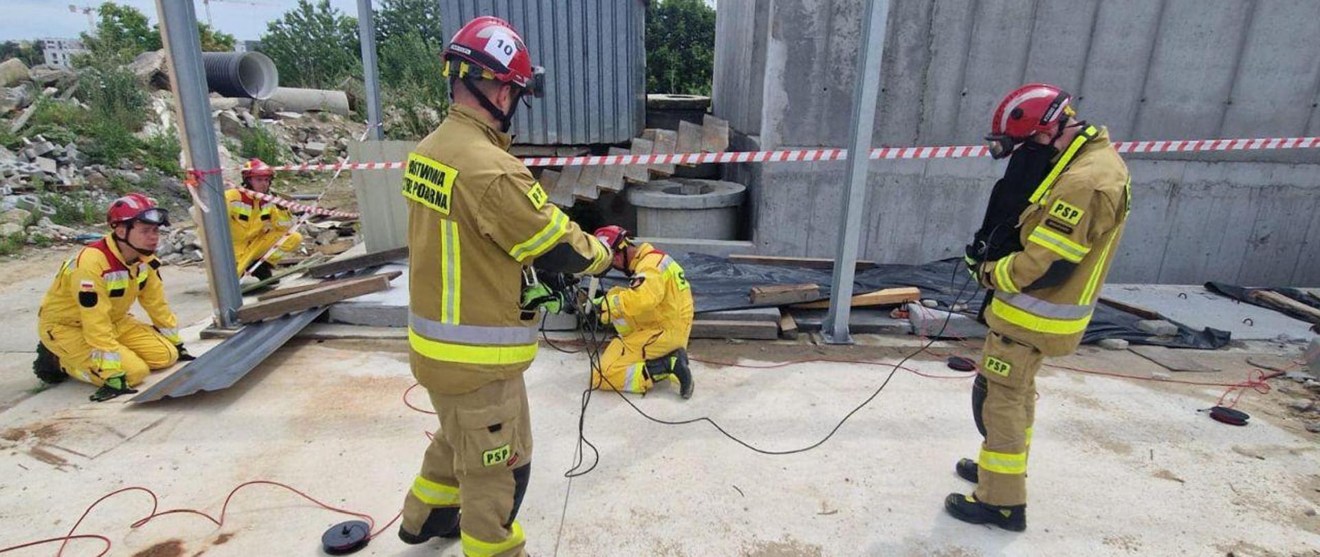 Strażacy są w momencie ćwiczeń. W tle symulowane gruzowisko. Strażacy ćwiczą użycie geofonu.