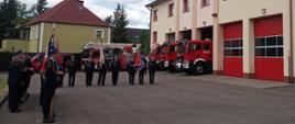 Poczty sztandarowe oraz strażacy, którzy stoją w dwuszeregu na placu Komendy Powiatowej PSP w Człuchowie. W tle samochody pożarnicze.