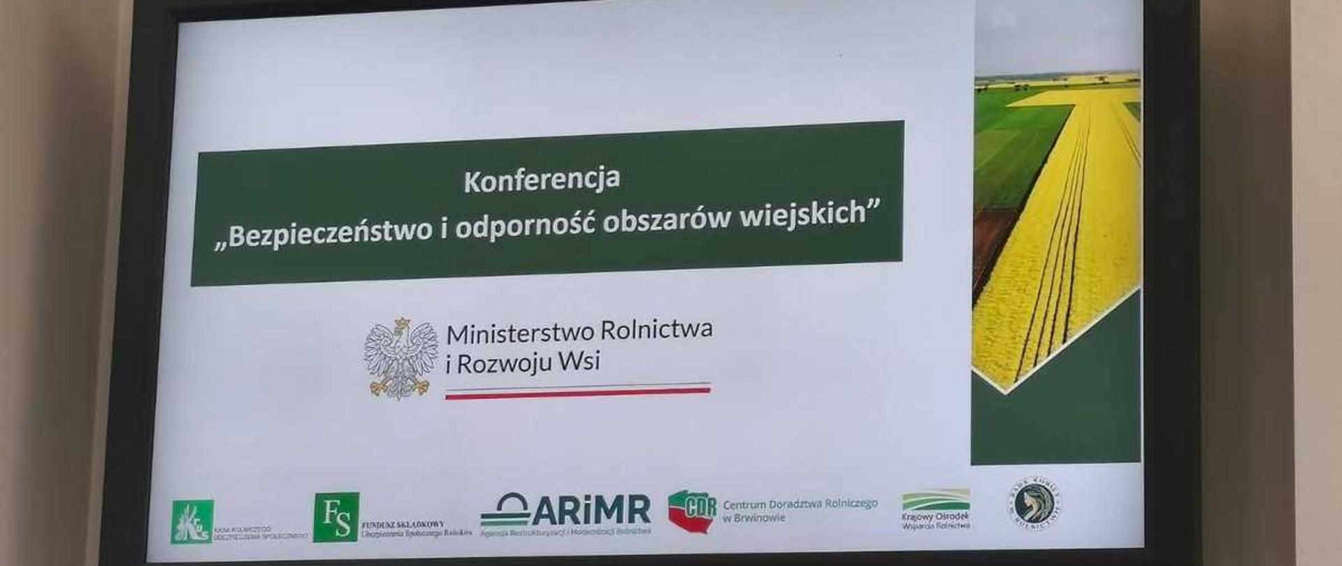Zdjęcie przedstawia duży, czarny ekran telewizyjny lub monitor marki NEC, na którym wyświetlany jest slajd tytułowy prezentacji konferencyjnej. Ekran wisi na jasnej, gładkiej ścianie.Treść slajduGłówny tytuł: W ciemnozielonej ramce na środku znajduje się biały napis: „Konferencja „Bezpieczeństwo i odporność obszarów wiejskich””.Organizator: Poniżej tytułu widnieje oficjalne logo z godłem Polski (orzeł w koronie) oraz podpis: „Ministerstwo Rolnictwa i Rozwoju Wsi”, podkreślony czerwoną linią.Grafika: Po prawej stronie slajdu umieszczono pionowe zdjęcie przedstawiające widok z góry na wiejski krajobraz – zielone i żółte pola uprawne rozdzielone prostymi liniami dróg.Dolny pasek z logotypami partnerówNa białym tle w dolnej części ekranu ułożono poziomo małe logotypy instytucji rolniczych:Zielony kwadrat Kasy Rolniczego Ubezpieczenia Społecznego (KRUS).Zielony kwadrat Funduszu Składkowego Ubezpieczenia Społecznego Rolników (FS).Niebiesko-czarne logo Agencji Restrukturyzacji i Modernizacji Rolnictwa (ARiMR).Czerwono-zielone logo Centrum Doradztwa Rolniczego w Brwinowie (CDR).Zielone logo Krajowego Ośrodka Wsparcia Rolnictwa (KOWR).Okrągłe, ciemne logo Rady Kobiet w Rolnictwie.