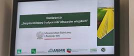 Zdjęcie przedstawia duży, czarny ekran telewizyjny lub monitor marki NEC, na którym wyświetlany jest slajd tytułowy prezentacji konferencyjnej. Ekran wisi na jasnej, gładkiej ścianie.Treść slajduGłówny tytuł: W ciemnozielonej ramce na środku znajduje się biały napis: „Konferencja „Bezpieczeństwo i odporność obszarów wiejskich””.Organizator: Poniżej tytułu widnieje oficjalne logo z godłem Polski (orzeł w koronie) oraz podpis: „Ministerstwo Rolnictwa i Rozwoju Wsi”, podkreślony czerwoną linią.Grafika: Po prawej stronie slajdu umieszczono pionowe zdjęcie przedstawiające widok z góry na wiejski krajobraz – zielone i żółte pola uprawne rozdzielone prostymi liniami dróg.Dolny pasek z logotypami partnerówNa białym tle w dolnej części ekranu ułożono poziomo małe logotypy instytucji rolniczych:Zielony kwadrat Kasy Rolniczego Ubezpieczenia Społecznego (KRUS).Zielony kwadrat Funduszu Składkowego Ubezpieczenia Społecznego Rolników (FS).Niebiesko-czarne logo Agencji Restrukturyzacji i Modernizacji Rolnictwa (ARiMR).Czerwono-zielone logo Centrum Doradztwa Rolniczego w Brwinowie (CDR).Zielone logo Krajowego Ośrodka Wsparcia Rolnictwa (KOWR).Okrągłe, ciemne logo Rady Kobiet w Rolnictwie.