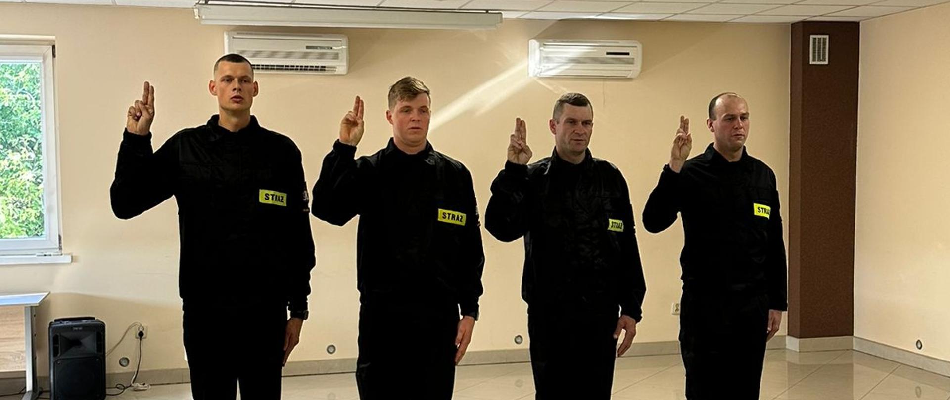 Nowo przyjęci strażacy w czarnych mundurach składają uroczyste ślubowanie, stojąc w szeregu z uniesioną dłonią.