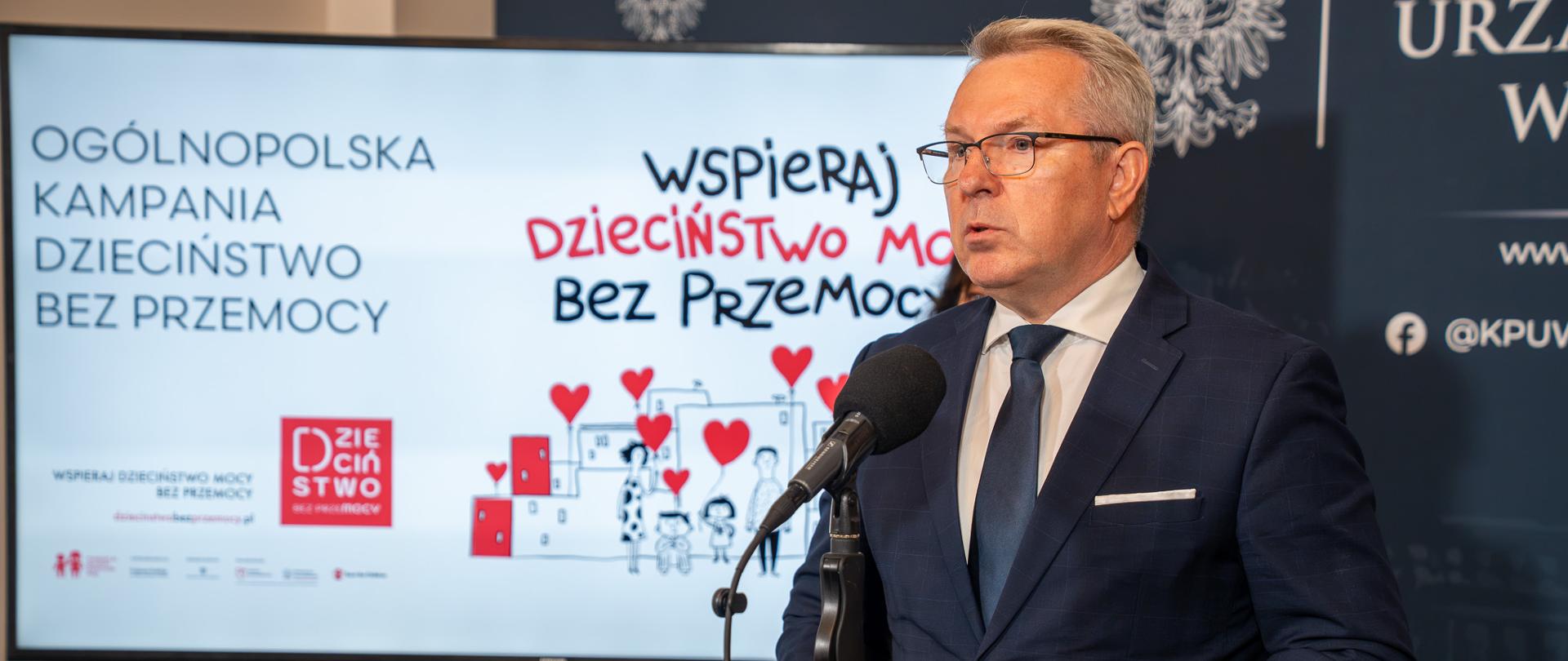 Wicewojewoda Piotr Hemmerling w ciemnym garniturze przemawia do mikrofonu, trzymając w rękach granatową teczkę. Po jego prawej stronie stoi ekran z grafiką promocyjną akcji. Na ekranie widnieje napis: „Ogólnopolska Kampania Dzieciństwo bez Przemocy” oraz „Wspieraj dzieciństwo bez przemocy”.
