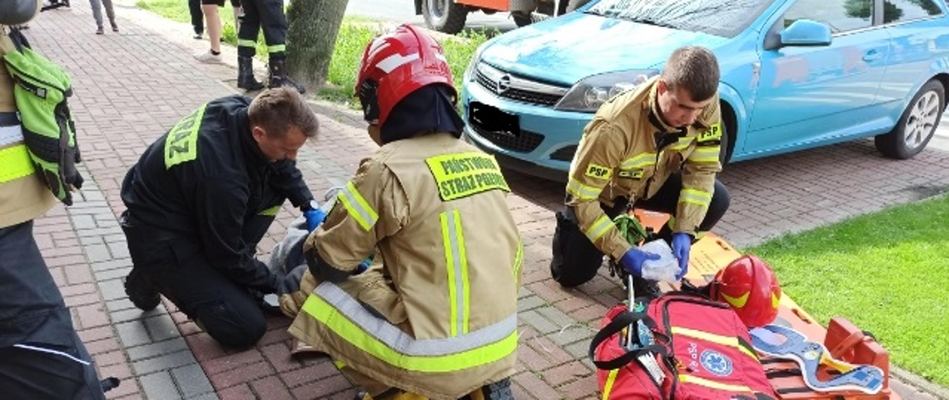 Widać strażaków udzielających pomocy medycznej osobie poszkodowanej.