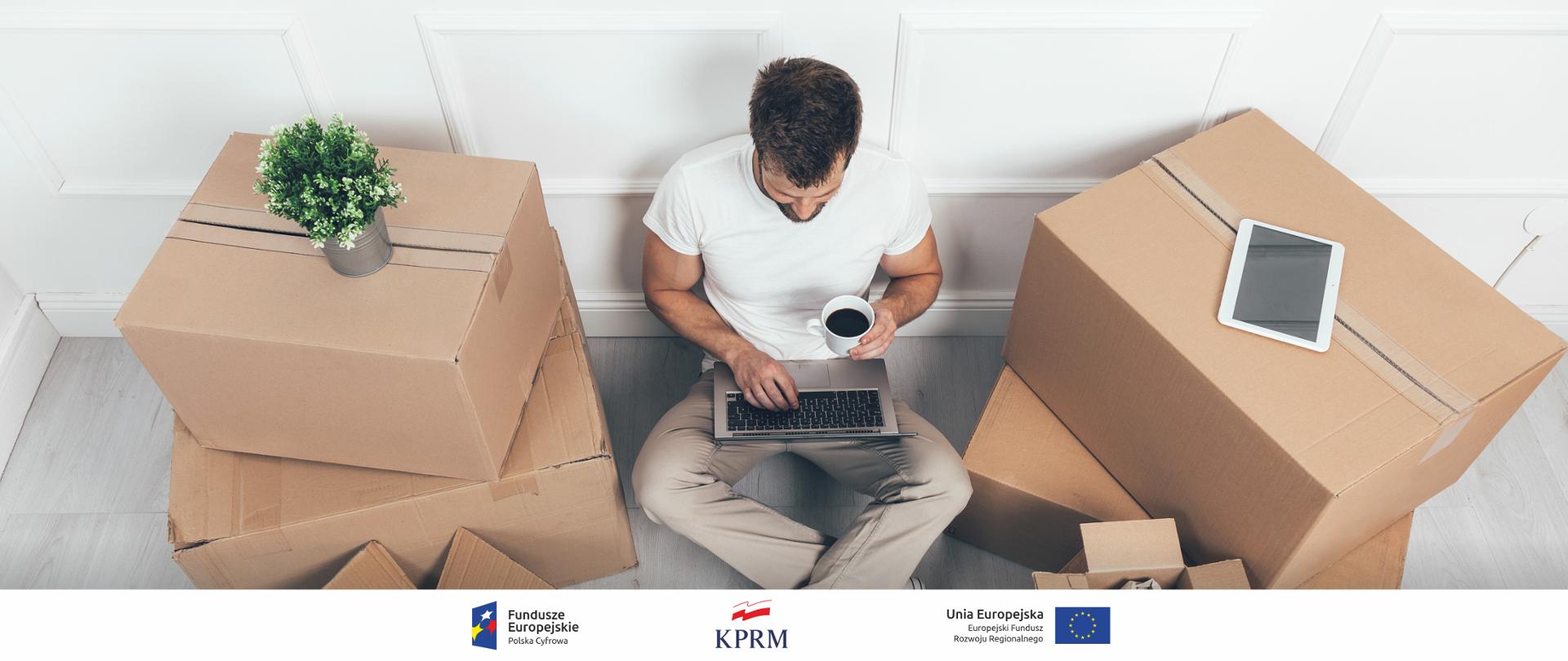 Zdjęcie, ujęcie od góry - mężczyzna siedzący na podłodze, w trakcie przeprowadzki, naokoło niego stoją pudełka. W jednym ręku trzyma kawę, drugą korzysta z laptopa.