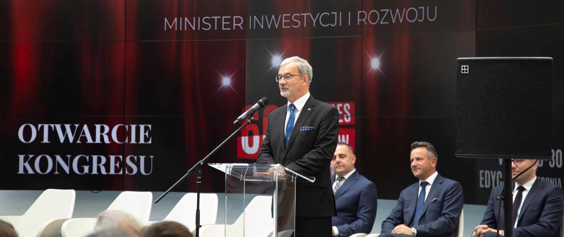 Minister Jerzy Kwieciński przemawia do mikrofonu stojąc przy przezroczystej mównicy. Za nim i przed nim siedzą osoby.