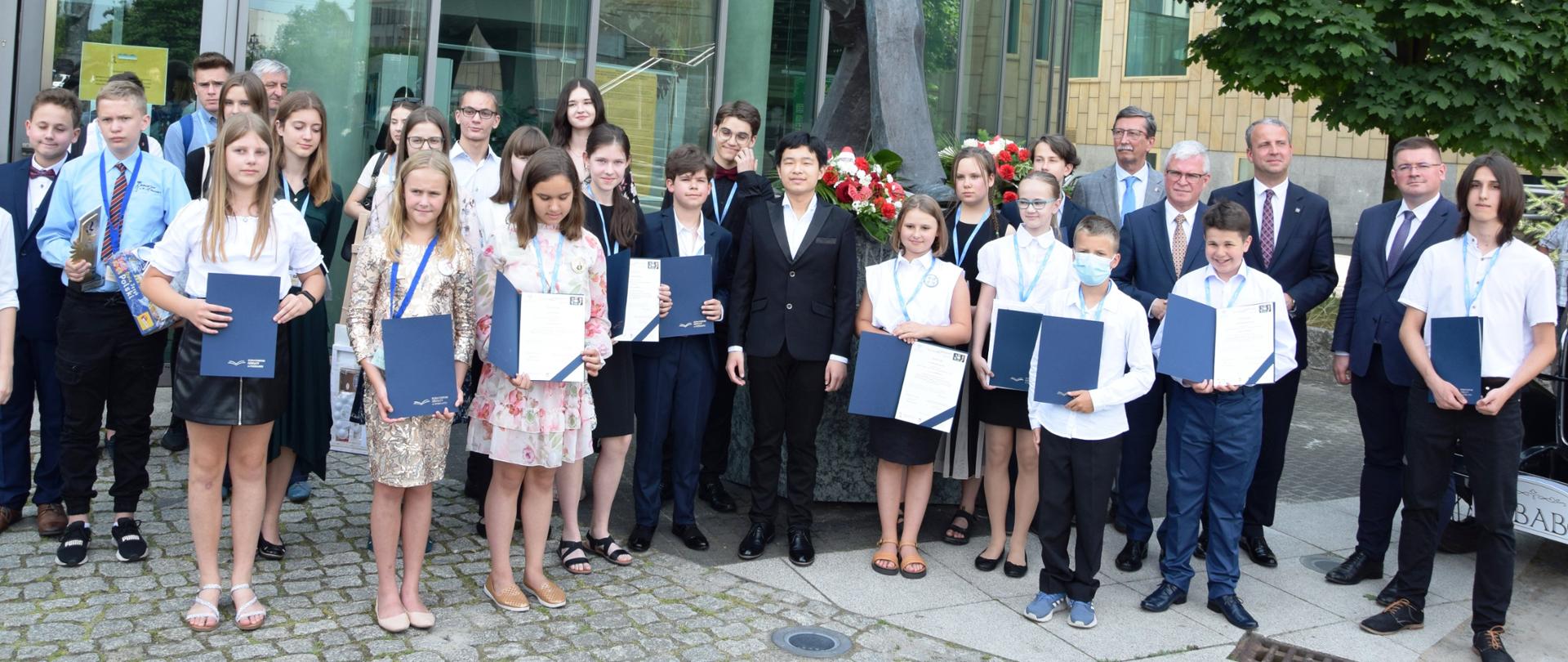 Grupa dzieci stojących przed budynkiem. Dzieci mają medale, w rękach trzymają teczki z dyplomami. Po prawej stronie wiceminister Rzymowski, profesor Jan Żaryn i przedstawiciele organizatorów przedsięwzięcia. 