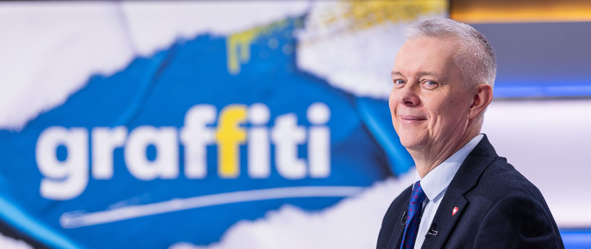 Na zdjęciu minister Tomasz Siemoniak (po prawej stronie) na tle wnętrza studnia telewizyjnego Polsat News. W tle po lewej stronie widać nazwę programu „Graffiti” (biało i żółte litery na niebieskim i białym tle). 