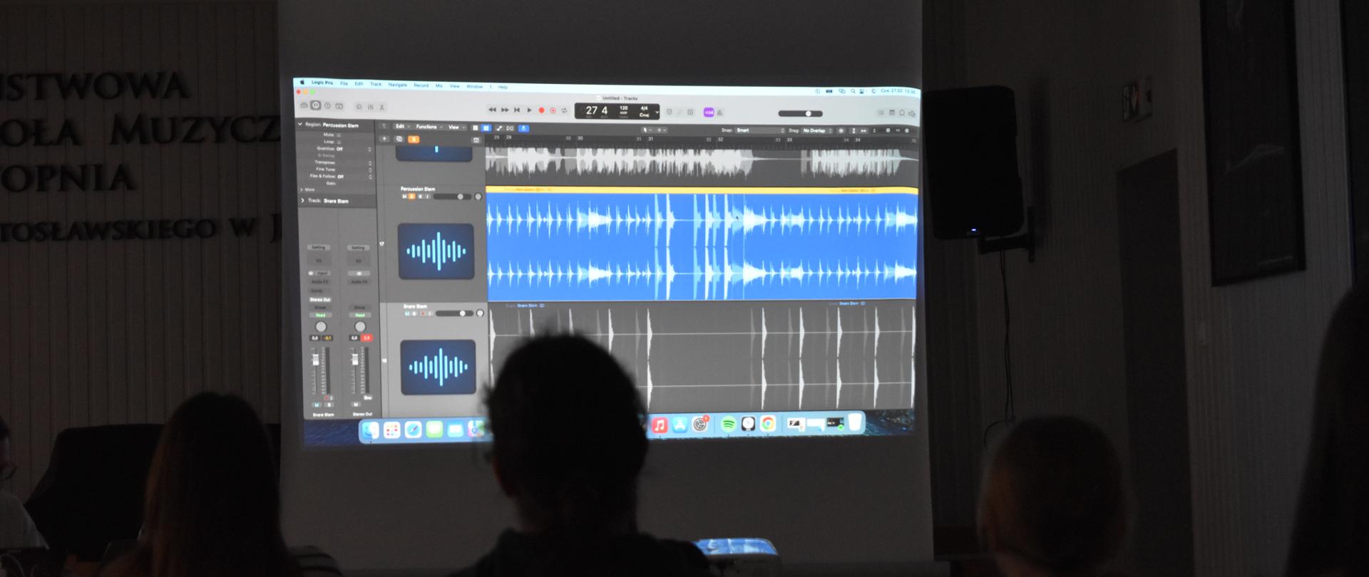 Na zdjęciu, w auli PSM widnieją uczniowie, którzy obserwują obraz z rzutnika wyświetlony na ekranie, który przedstawia cyfrowy zapis ścieżki audio. Kolorystyka zdjęcia jest szaro-biało-niebiesko-żółta.