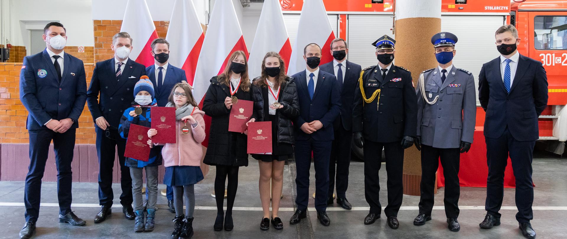 Ośmiu mężczyzn i czworo dzieci stoi na tle flag i wozu strażackiego. Dzieci mają w rękach medale i upominki. 