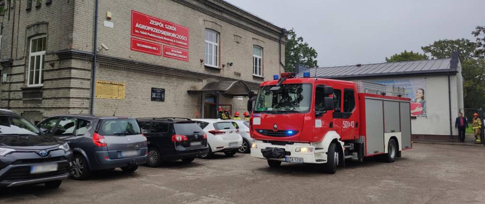 Zdjęcie przedstawia strażaków w ubraniach specjalnych podczas wchodzenia do budynku szkoły z linią gaśniczą w celu ugaszenia pozorowanego w ramach próbnej ewakuacji pożaru w jednym z pomieszczeń szkoły. Na zdjęciu widać część budynku szkoły, zaparkowane przy szkole cywilne samochody osobowe oraz na pierwszym planie średni samochód ratowniczo-gaśniczy stojący na środku parkingu z włączonymi światłami mijania i niebieskimi światłami błyskowymi. Zdjęcie w pochmurny dzień wykonano przed budynkiem Zespołu Szkół Agroprzedsiębiorczości w Zambrowie. 