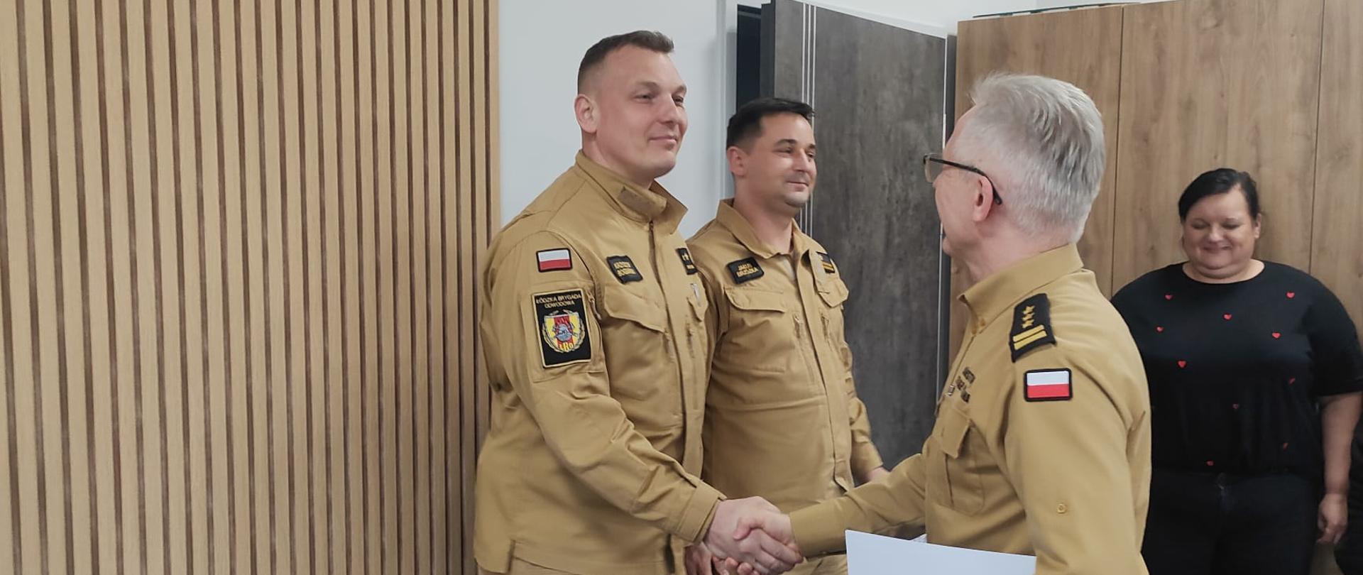 Komendant Powiatowy Państwowej Straży Pożarnej w Brzezinach starszy brygadier Mariusz Kaczmarski składa podziękowania kapitanowi Krzysztofowi Bogdańskiemu za działania
w zakresie udzielenia pomocy nieprzytomnemu mężczyźnie, u którego doszło do nagłego zatrzymania krążenia. W lewym ręku komendant trzyma teczkę z listem gratulacyjnym prawą ręką gratuluje kapitanowi. Okoliczności mają miejsce w gabinecie Komendanta Powiatowego.
