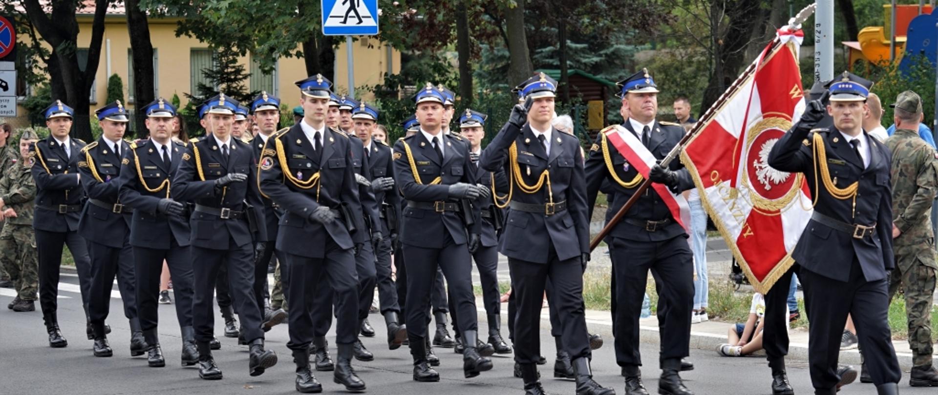 strażacka kompania reprezentacyjna maszeruje po ulicy, prowadzi ich poczet sztandarowy przed pocztem idzie salutujący strażaka
