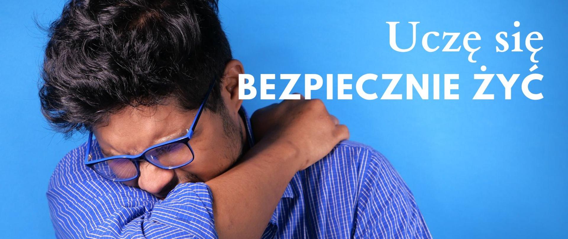 Mężczyzna zasłaniający twarz łokciem w czasie kichania. Napis Uczę się bezpiecznie żyć.