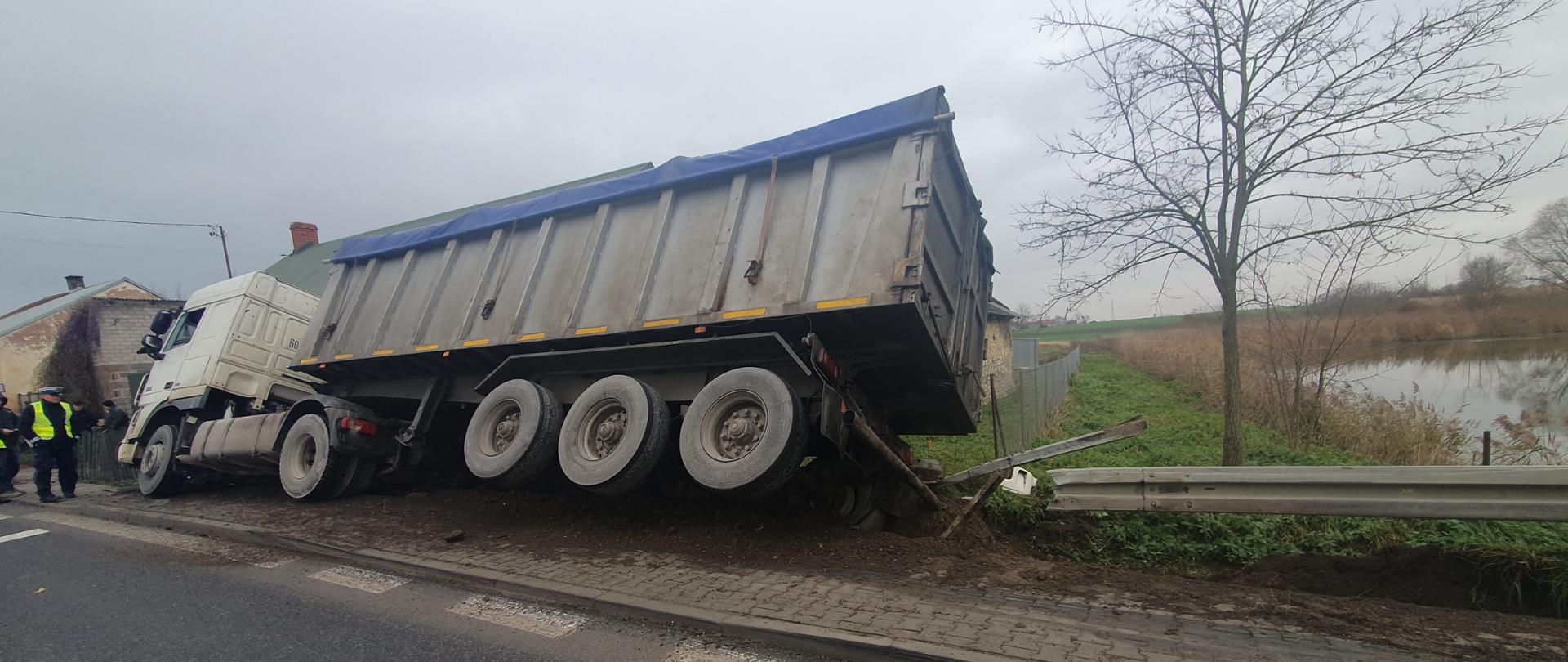 Na zdjęciu widać ciągnik siodłowy z naczepą przechylony na budynku jednorodzinnym.