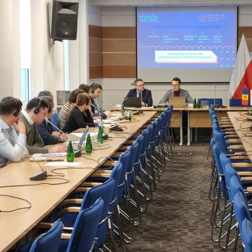 Sala konferencyjna ze stołami przy których siedzą ludzie z laptopami i słuchawkami. Wyświetlana jest prezentacja po angielsku, widać flagi Polski, Unii Europejskiej i Mołdawii