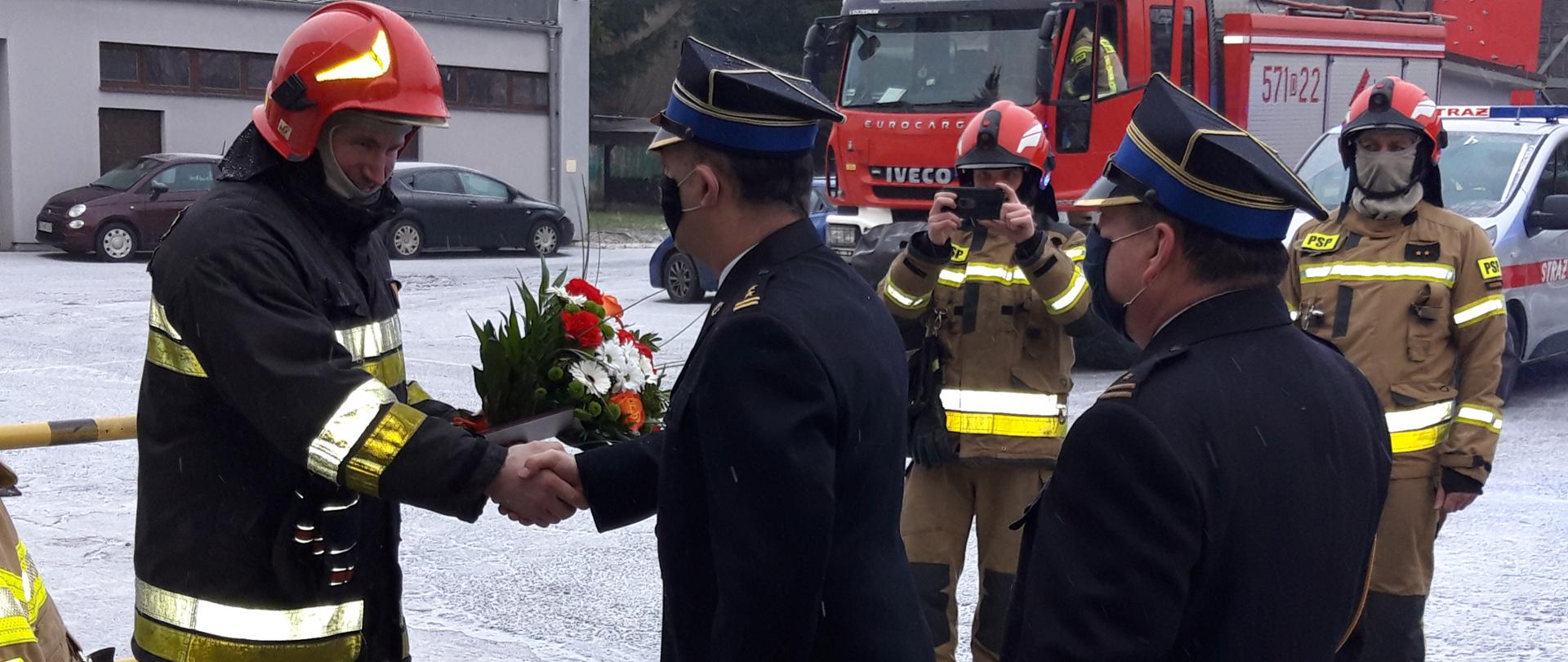 Na zdjęciu Komendant Powiatowy w mundurze galowym wręczający kwiaty strażakowi, który odchodzi na emeryturę.