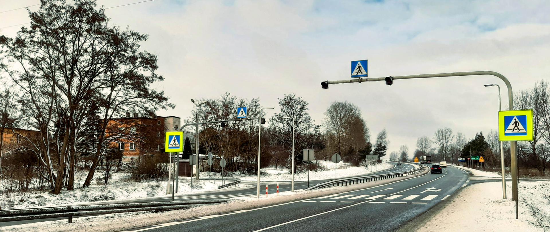 Fotografia z poziomu terenu przejścia dla pieszych przez dwujezdniową drogę w aurze zimowej.
Przejście dla pieszych posiada wydzieloną wyspę separacyjną pomiędzy jezdniami. Nad przejściami zamontowano na wysięgnikach pulsujące światła ostrzegawcze. W rejonie przejścia dla pieszych znajdują się również zjazdy i wyjazdy na drogę krajową. W otoczeniu drogi widoczna zabudowa jednorodzinna.
