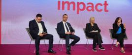 Na oświetlonym podwyższeniu z przodu sali na krzesłach siedzą cztery osoby, drugi od lewej minister Kulasek, za nimi na wielkim ekranie napis Impact.