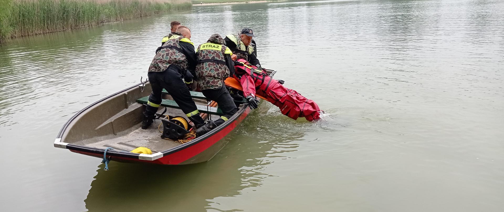 Zdjęcie wykonane z brzegu, na wodzie pływa łódź strażacka. W łodzi jest czterech ratowników, którzy wyciągają z wody strażaka w czerwonym skafandrze. 