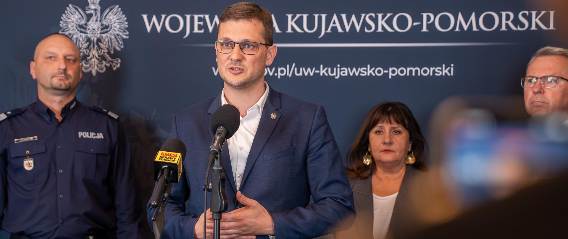 cztery osoby stoją na tle ścianki, przed jedną z nich stoją dwa mikrofony 
