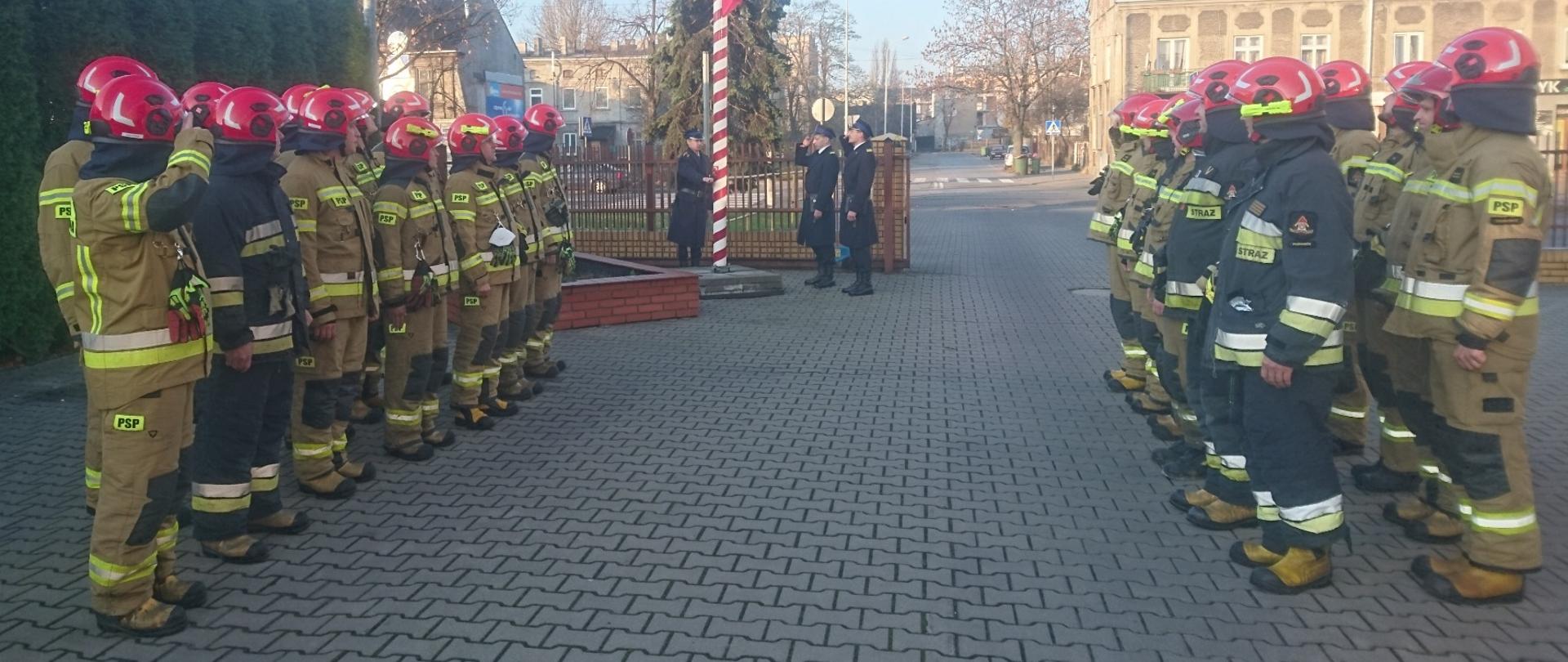 Na zdjęciu po lewej i prawej stronie kadru stoją strażacy dwóch zmian służbowych ubrani w ubrania specjalne, pomiędzy nimi przy maszcie znajduje się poczet flagowy ubrany w mundury wyjściowe.