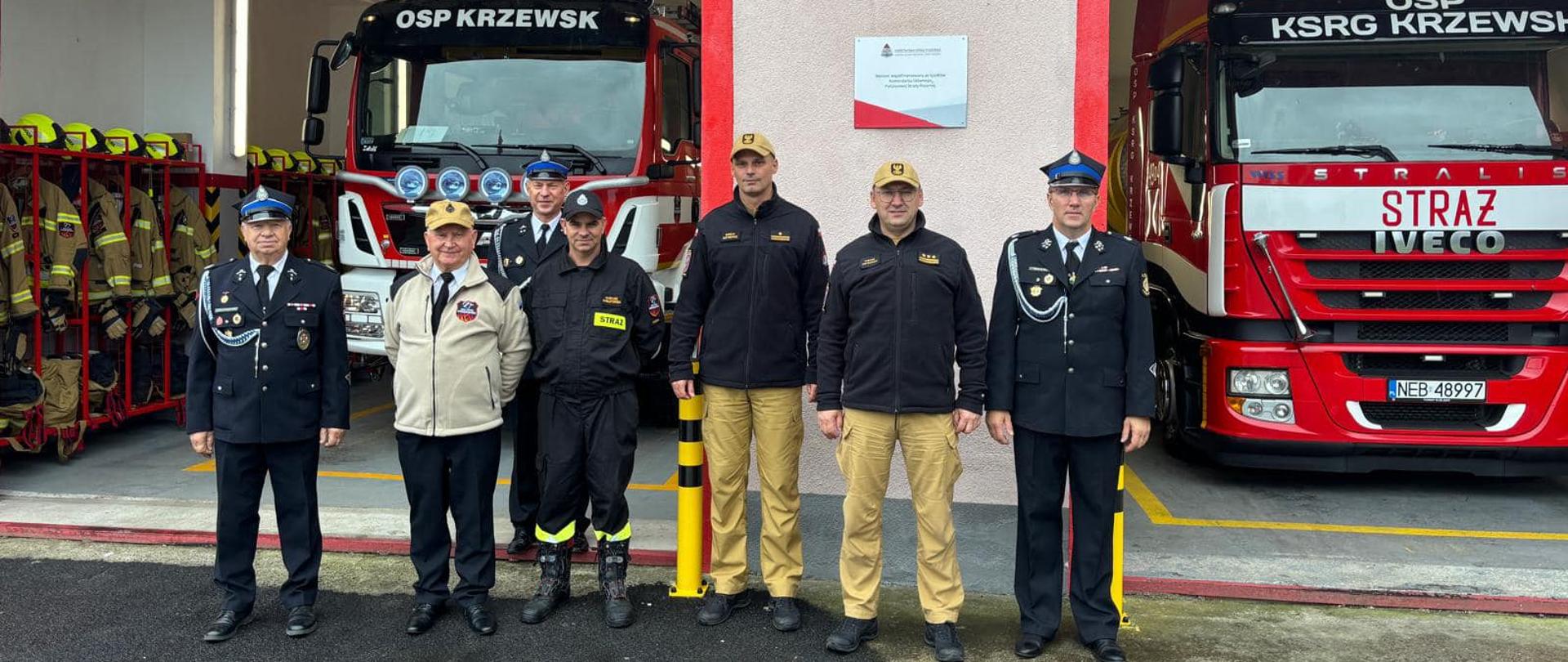 Na zdjęciu znajduje się Zastępca Warmińsko - Mazurskiego Komendanta Wojewódzkiego PSP, Komendant Miejski PSP w Elblągu i strażacy ochotnicy z OSP w Krzewsku. Osoby stoją na tle budynku remizy i dwóch pojazdów gaśniczych