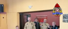 Na zdjęciu widać spotkanie w sali Domu Kultury, podczas którego strażacy z Państwowej Straży Pożarnej w Gryficach prowadzą prelekcję dla mieszkańców. Dwóch funkcjonariuszy w beżowych mundurach stoi przed uczestnikami, a jeden z nich trzyma w rękach kartki z materiałami informacyjnymi. Obok nich znajduje się kilku mężczyzn, w tym fotograf wykonujący zdjęcia oraz osoba w garniturze siedząca przy stoliku. W tle widoczny jest baner Komendy Powiatowej PSP w Gryficach z logo straży pożarnej. W prawym górnym rogu zdjęcia umieszczone jest logo Państwowej Straży Pożarnej z napisem „Gryfice”.
