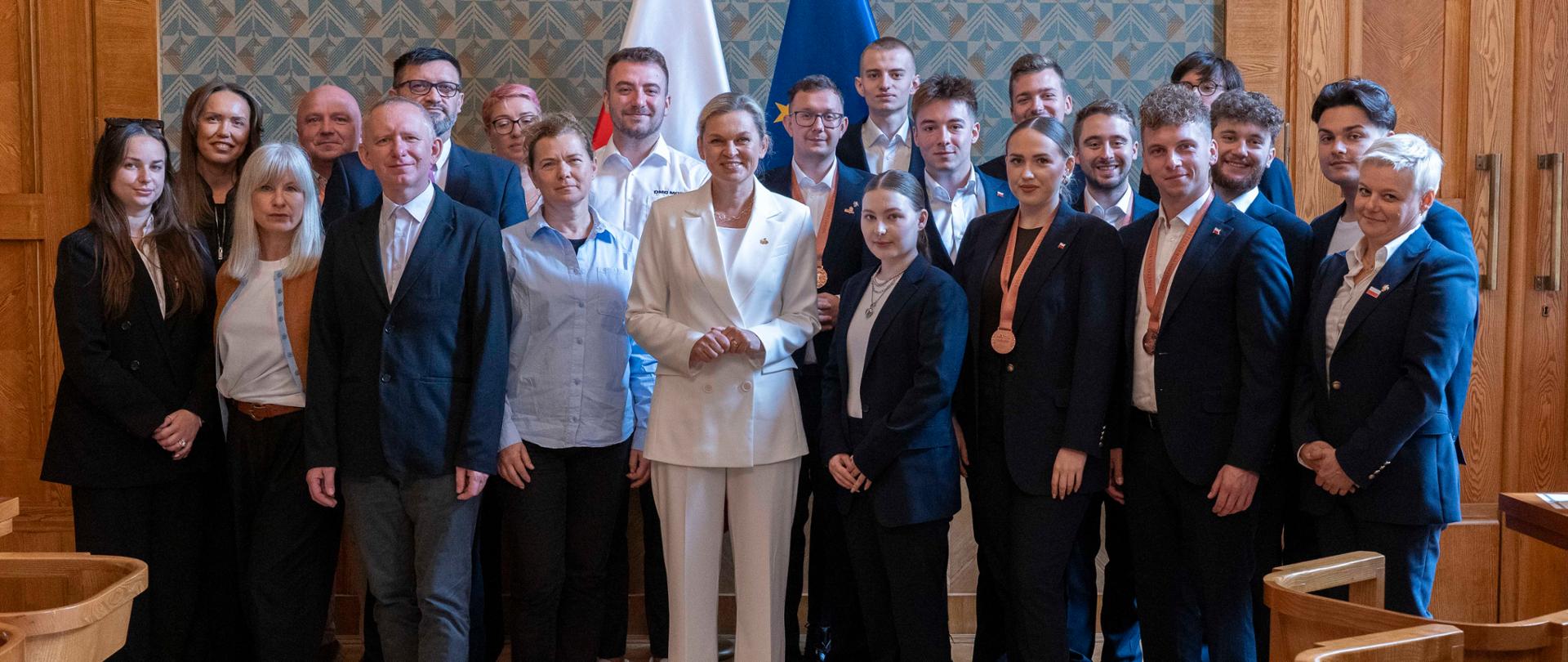 Duża grupa osób, zarówno mężczyzn, jak i kobiet, w różnym wieku, pozuje do zdjęcia w sali z drewnianymi panelami i tapetą. Kobieta w białym garniturze stoi na środku. W tle widoczne są polskie i europejskie flagi.