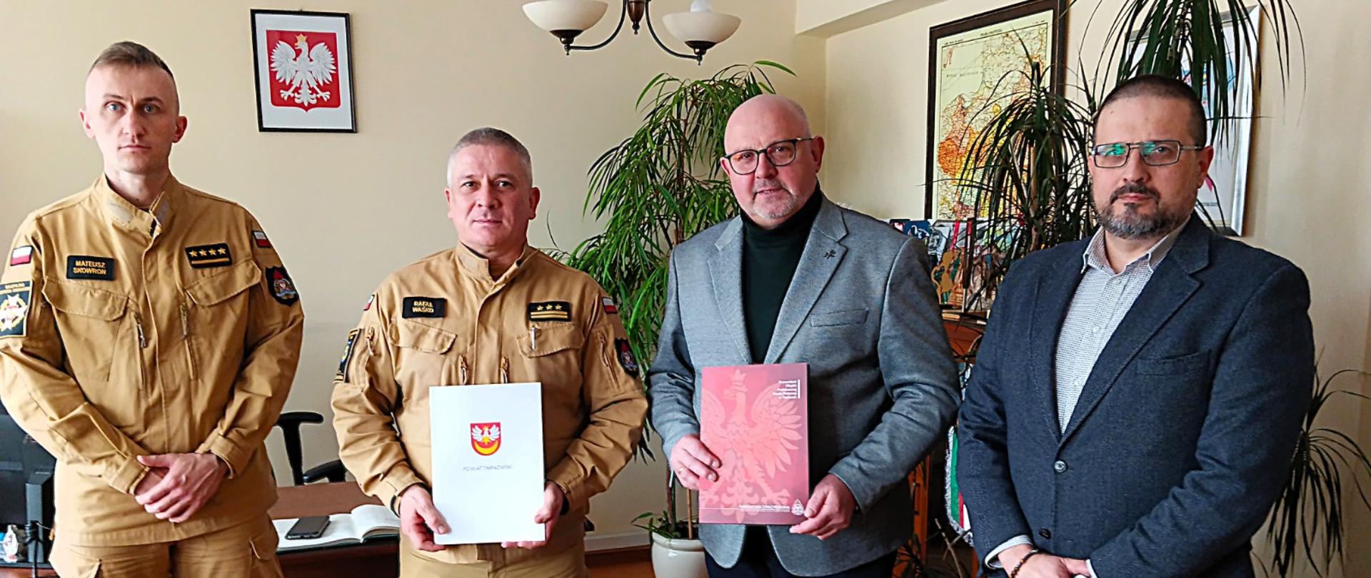 Strażacy w mundurach piaskowych stają w rzędzie z dwoma mężczyznami. W rękach trzymają teczki z podpisanymi porozumieniami. W tle żółta ściana z godłem Polski