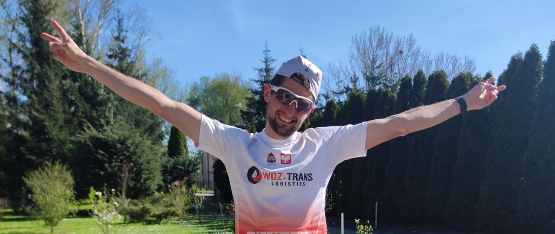 mężczyzna w w okularach słonecznych i czapeczce stoi z rozłożonymi rękoma i uśmiechem