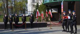 Trzech strażaków maszeruje. Jeden z nich niesie na rękach biało-czerwona flagę. Po lewej stronie stoi pięciu strażaków. W tle widać ustawione przed budynkiem biało czerwone flagi. Za nimi jasny budynek i zielone drzewa. 