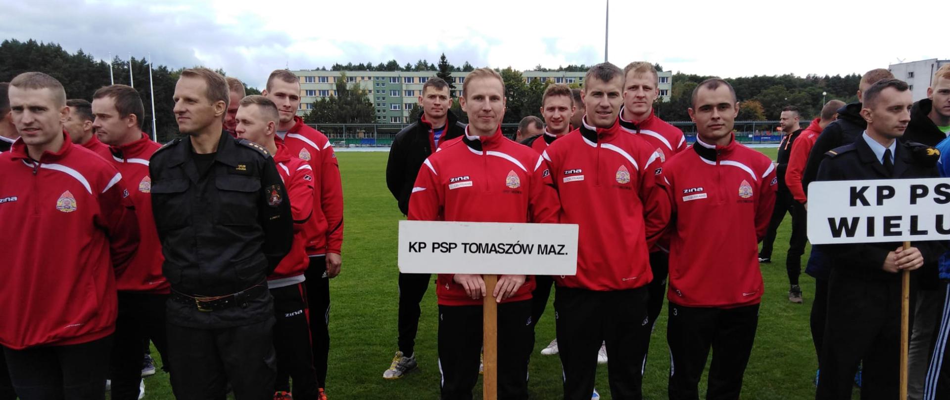 Mężczyźni w strojach sportowych stoją na zielonej trawie. Dwóch trzyma tabliczki z napisami. w tle widać drzewa i bloki. 