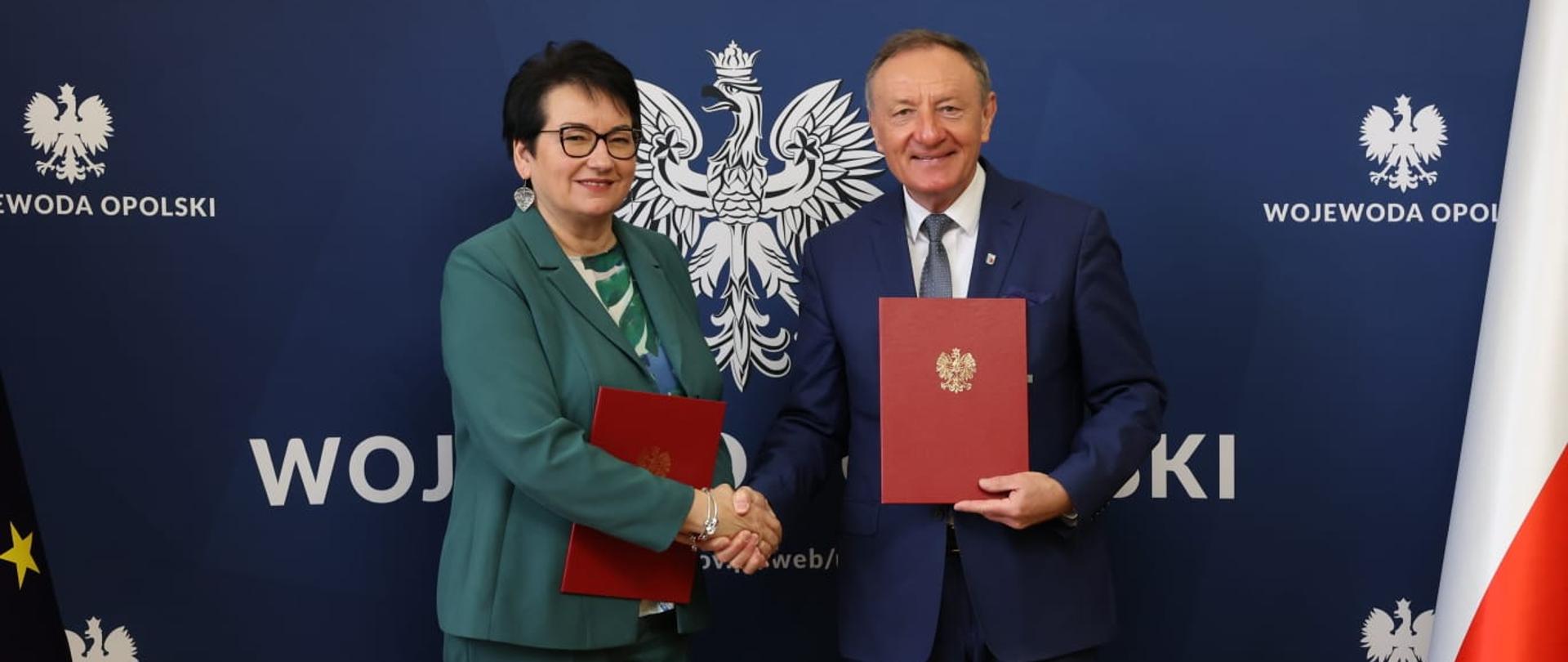 Wojewoda Opolski Monika Jurek z Burmistrzem Olesna Sylwestrem Lewickim. Na zdjęciu dają sobie uścisk dłoni. Za nimi widać granatowy baner z napisami "Wojewoda Opolski". 