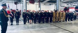 Wnętrze garażu. Na pierwszym planie po lewej stronie zdjęcia stoi dowódca uroczystości – strażak PSP w czarnym mundurze służbowym. W tle widoczni inni strażacy PSP w czarnych mundurach wyjściowych oraz piaskowych mundurach służbowych.