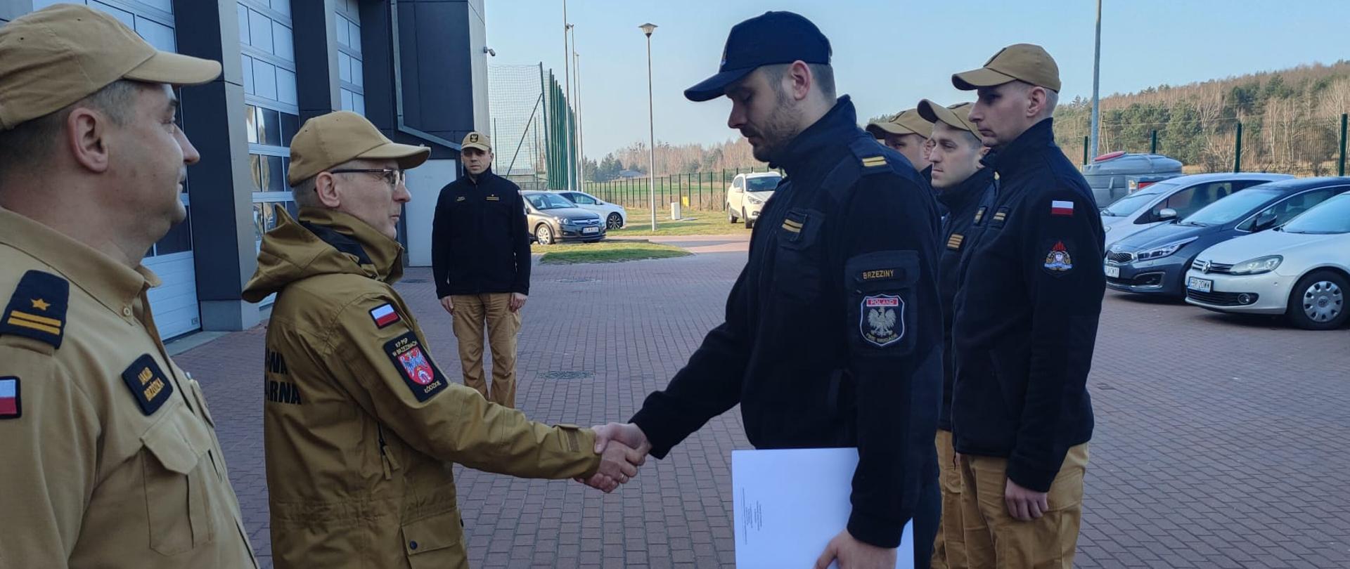 Komendant Powiatowy Państwowej Straży Pożarnej w Brzezinach starszy brygadier Mariusz Kaczmarski podczas zmiany służby składa podziękowania sekcyjnemu Damianowi Królowi podczas wypadku drogowego z udziałem trzech pojazdów, w tym nieoznakowanego radiowozu poruszającego się na sygnałach świetlnych i dźwiękowych. W wyniku zdarzenia cztery osoby zostały poszkodowane i wymagały udzielenia medycznych czynności ratunkowych. Komendant gratuluje funkcjonariuszowi poprzez uścisk dłoni. Wyróżniony funkcjonariusz w lewym ręku trzyma teczkę z listem gratulacyjnym wręczoną przez komendanta. Okoliczności mają miejsce na placu komendy podczas zmiany służby zgodnie z ceremoniałem pożarniczym. Obok komendanta asystuje Zastępca Komendanta Powiatowego Państwowej Straży Pożarnej w Brzezinach młodszy brygadier Jakub Brzózka.