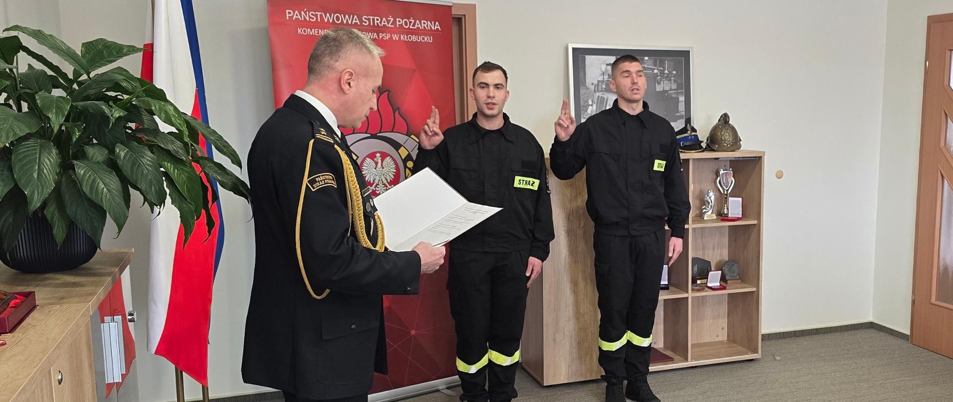 Uroczystość w pomieszczeniu biurowym Państwowej Straży Pożarnej. Dwóch strażaków w czarnych mundurach stoi wyprostowanych z uniesioną prawą ręką, składając ślubowanie. Przed nimi oficer w galowym mundurze czyta dokument. W tle widoczna flaga Polski, baner PSP oraz regał z hełmem i pucharami; z prawej strony drzwi i fragment sylwetki kolejnego funkcjonariusza.