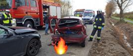Widok na tył jedno z rozbitych aut. Auto ma wgnieciony zderzak i klapę. Po prawej stronie stoi strażak a po lewej widać auto gaśnicze.