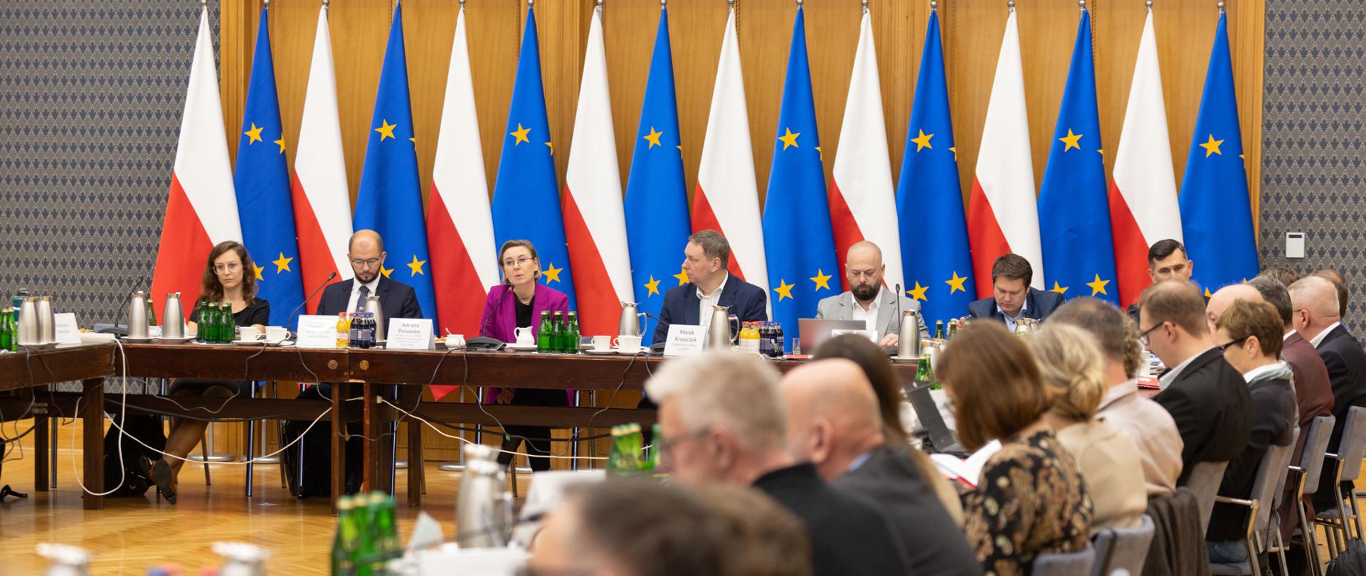 11.09.2025 r. Kancelaria Prezesa Rady Ministrów. Posiedzenie Rady Działalności Pożytku Publicznego, na zdjęciu uczestnicy wydarzenia siedzący za stołem konferencyjnym. W tle flagi Polski i Unii Europejskiej. Na pierwszym planie rozmyte sylwetki uczestników siedzących z drugiej strony stołu, tyłem do obiektywu. Na zdjęciu, widoczni od lewej: Adela Gąsiorowska, Zastępczyni dyrektora Narodowego Instytutu Wolności – Centrum Rozwoju Społeczeństwa Obywatelskiego, Michał Braun, Dyrektor Narodowego Instytutu Wolności – Centrum Rozwoju Społeczeństwa Obywatelskiego, Adriana Porowska, Sekretarz Stanu w KPRM, Wiceprzewodnicząca Komitetu ds. Pożytku Publicznego, Marek Krawczyk, Podsekretarz Stanu w KPRM, Maciej Kunysz, Stowarzyszenie EKOSKOP, Współprzewodniczący Rady oraz Łukasz Jachimowicz, Dyrektor Departamentu Społeczeństwa Obywatelskiego KPRM.