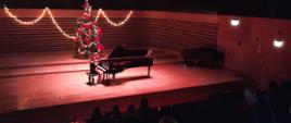 Dziewczynka gra na fortepianie na estradzie sali koncertowej, z przodu widać od tyłu widownię zapełnioną publicznością.