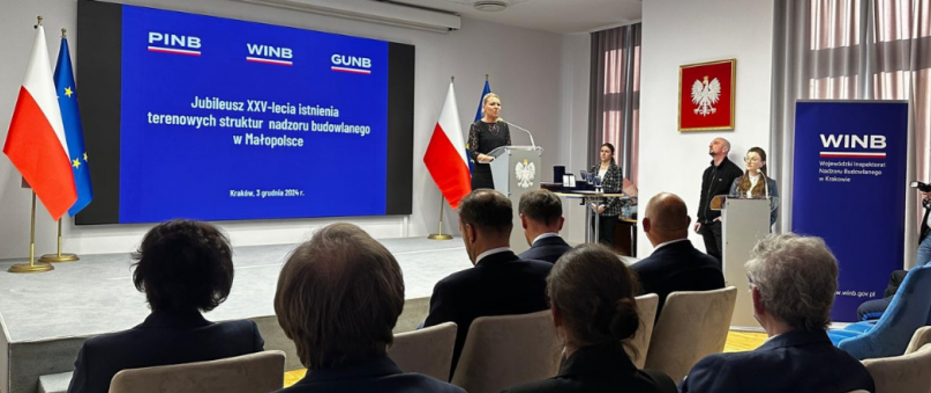 Sala konferencyjna podczas jubileuszu XXV-lecia nadzoru budowlanego w Małopolsce; uczestnicy słuchają przemówienia przy mównicy, w tle polska i unijna flaga oraz ekran z informacją o wydarzeniu.