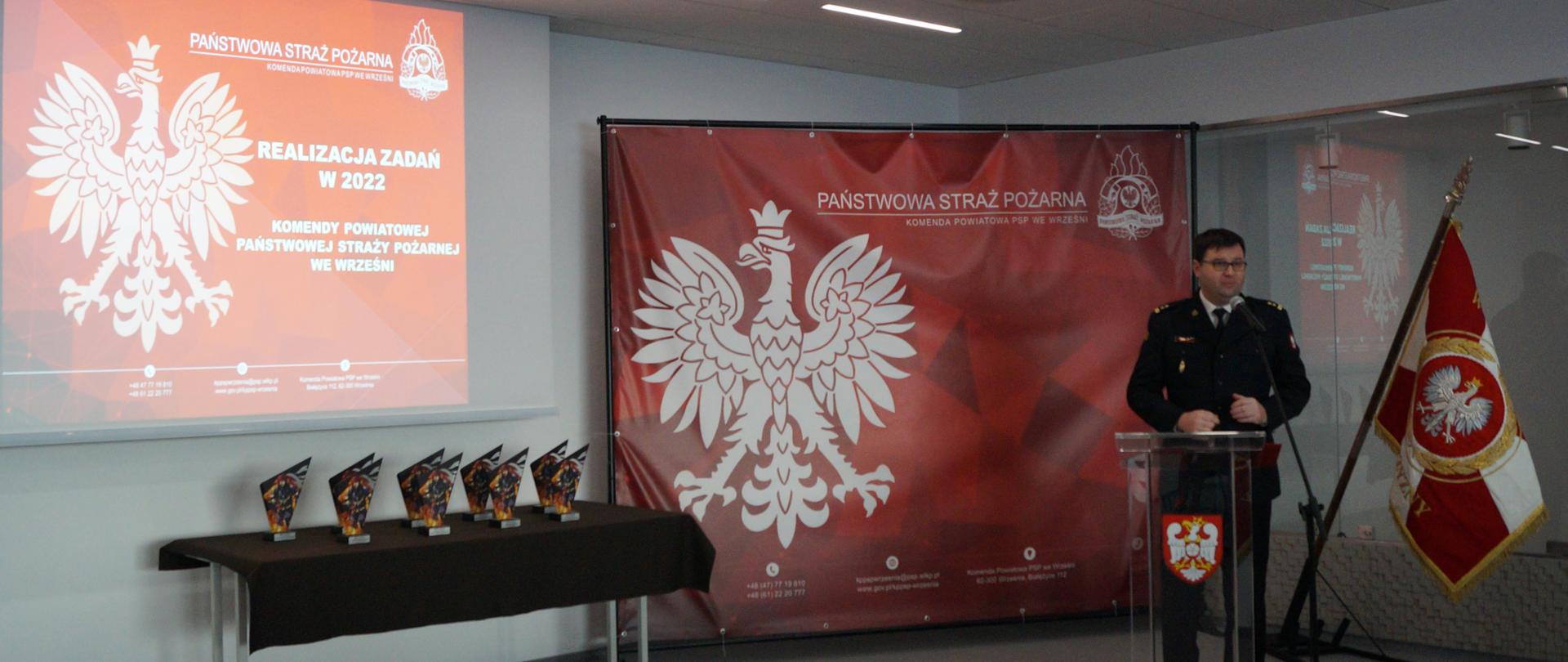 strażak w mundurze wyjściowym stoi przy mównicy, obok sztandar jednostki odwrócony biało czerwoną stroną, za strażakiem czerwona ścianka tekstylna z orłem i logo PSP, w tle ekran z prezentacją zawierającą orla i napisy, poniżej ekranu stół ze statuetkami