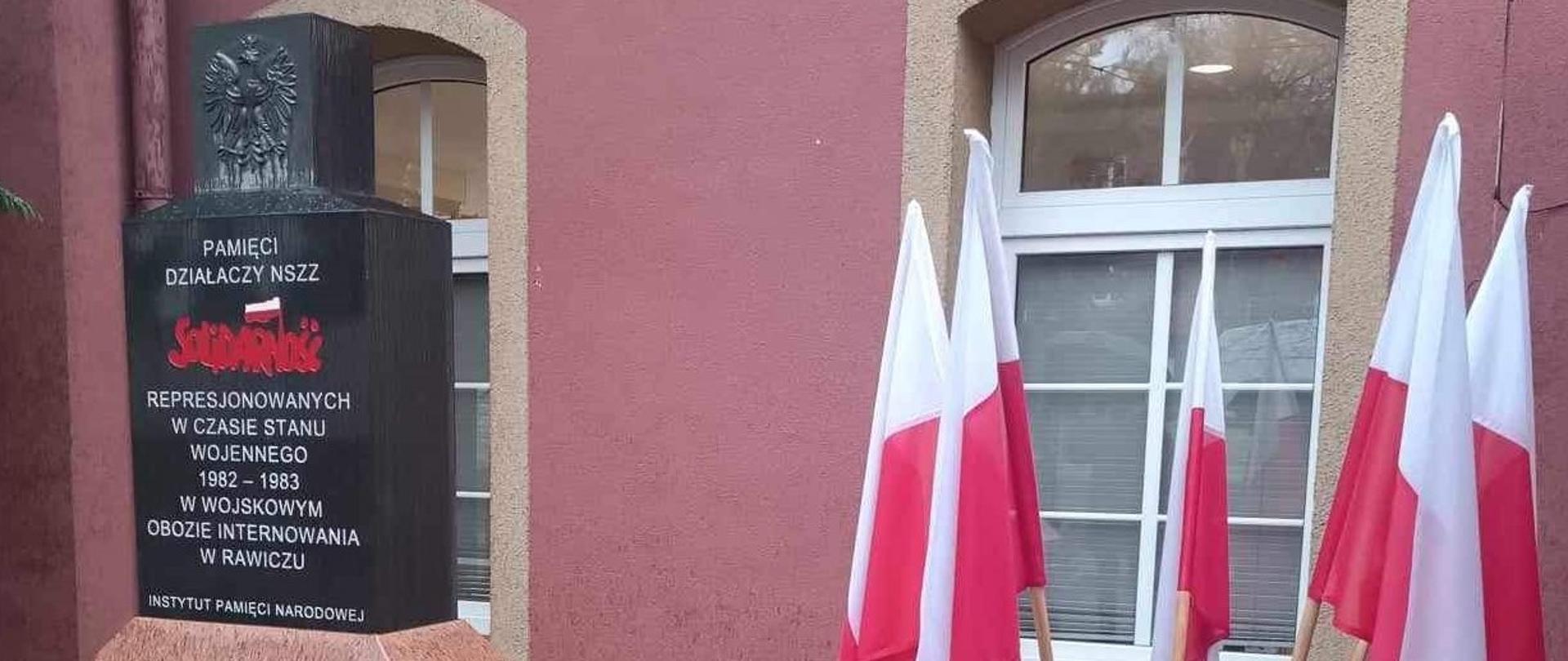 Na tle ściany budynku znajduje się pomnik - bryła w kolorze czarnym. Na jej szczycie umieszczone jest godło RP, a w centralnej części napis: Pamięci działaczy NSZZ Solidarność represjonowanych w czasie stanu wojennego 1982-1983 w Wojskowym Obozie Internowania w Rawiczu. Instytut Pamięci Narodowej. Z prawej strony stoi stojak z flagami biało-czerwonymi.