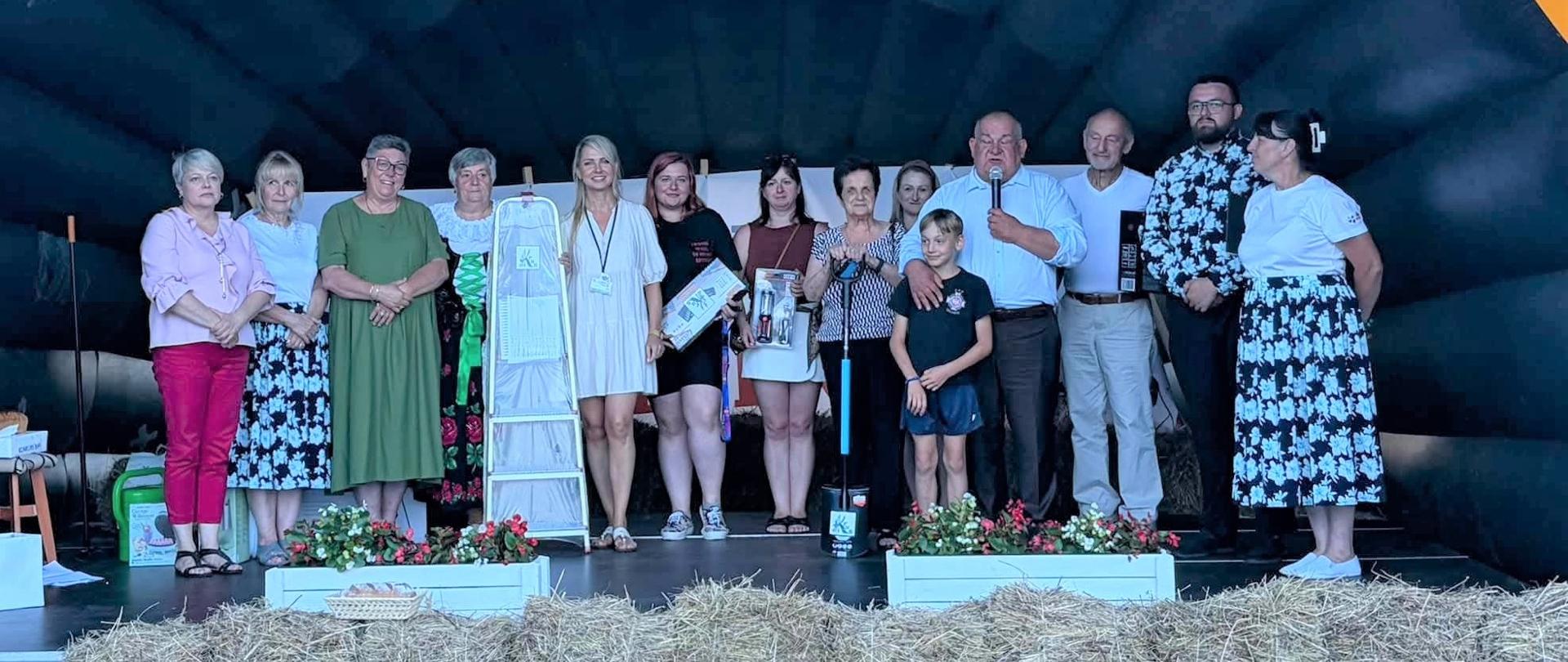 Na scenie znajduje się grupa osób-są to laureaci konkursu, który w rękach trzymają wygrane nagrody.